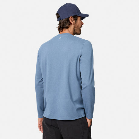 [Model is wearing size M.] A man wearing a heathered blue long-sleeve crewneck shirt, a navy blue baseball cap, and black pants, shown from a rear three-quarter view against a light gray studio background.