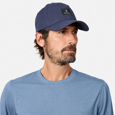 [Model is wearing size M.] A man wearing a navy blue baseball cap with a black rectangular logo patch and a heathered blue crewneck shirt, photographed against a clean light gray background.