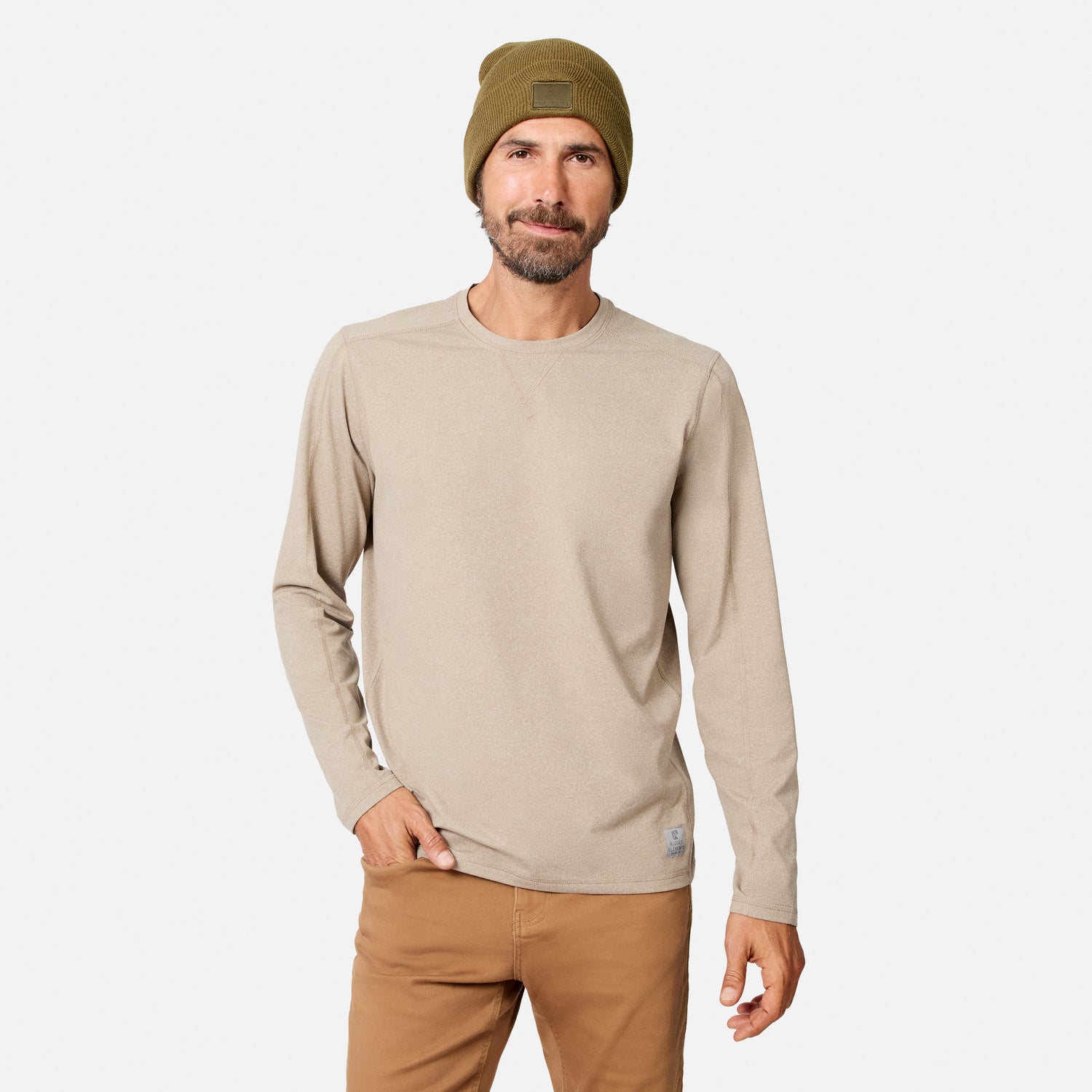 [Model is wearing size M.] A man wearing a light beige long-sleeve crewneck shirt, brown pants, and an olive green beanie, standing against a light gray background.