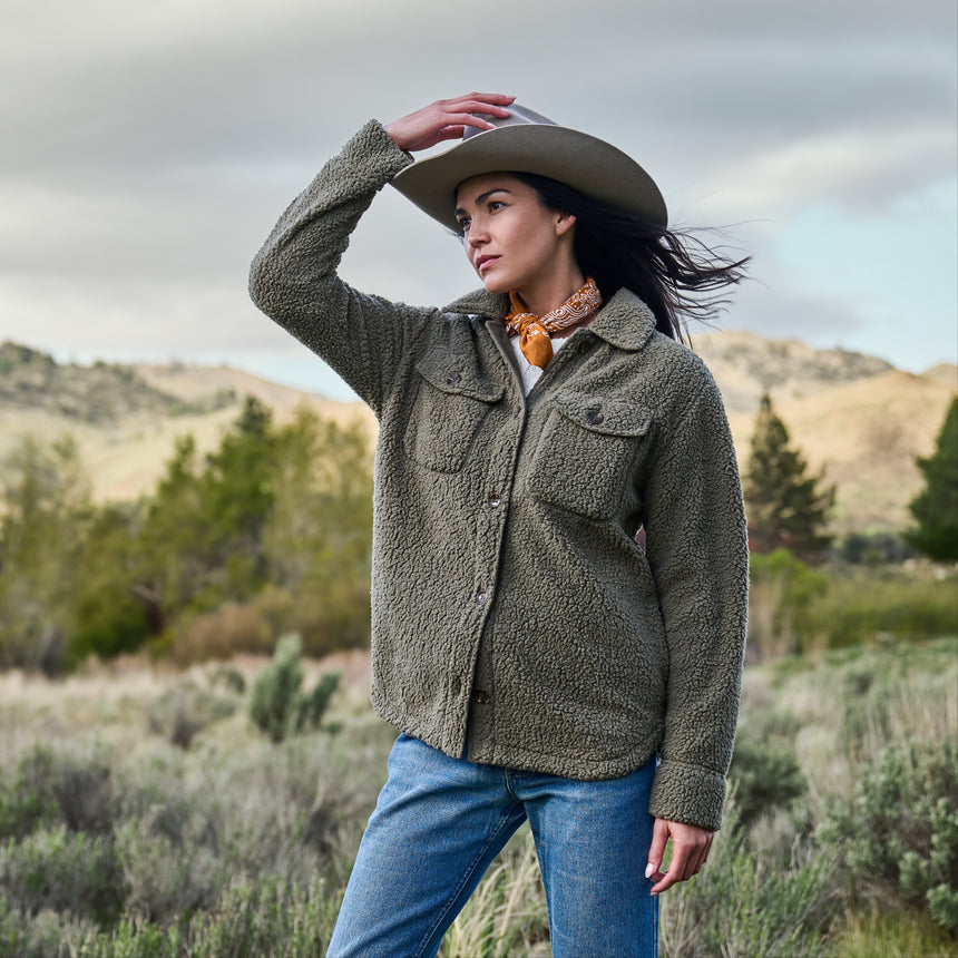 Woman in an olive sherpa shirt jacket holding her hat in the wind