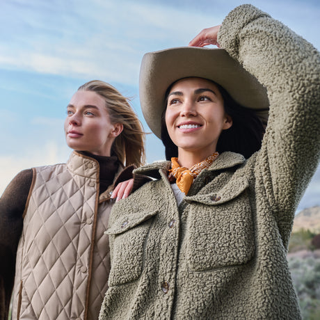 Two women outdoors under a blue sky, one wearing an olive green sherpa fleece button-up jacket with a cowboy hat and orange neck scarf, and the other wearing a beige quilted vest.