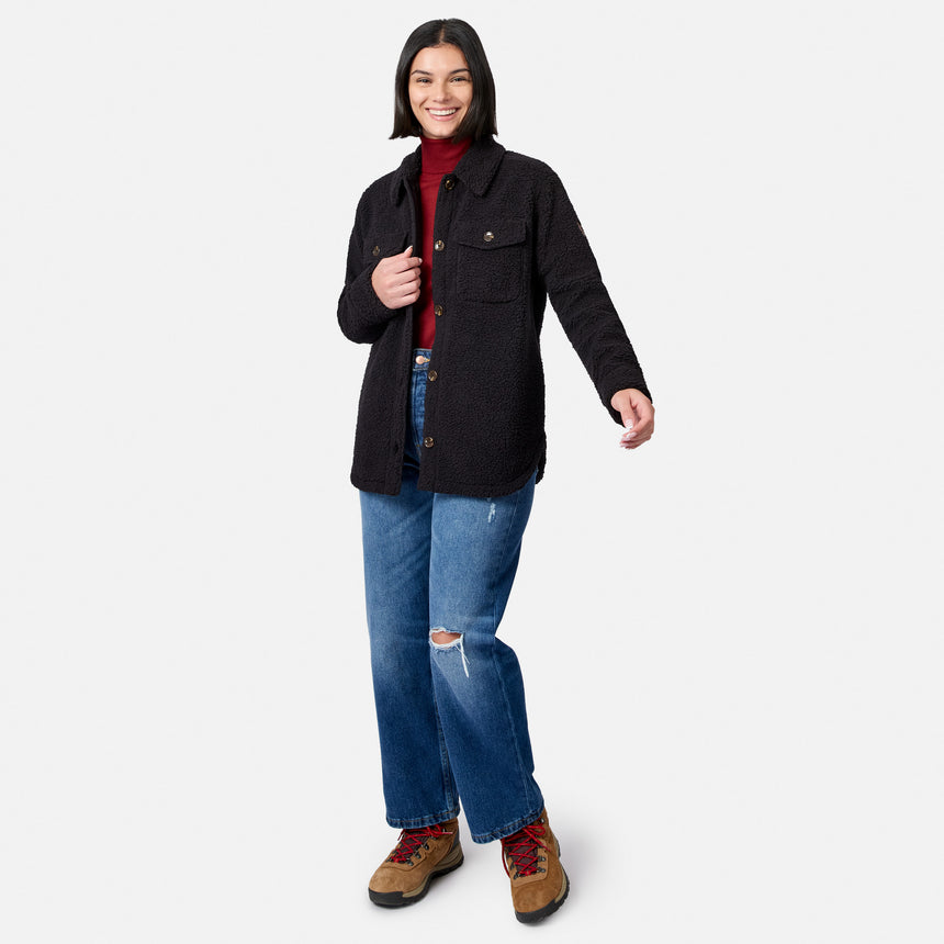 A smiling woman stands against a white background wearing the Free Country Women’s Sherpa Shacket, styled with a red turtleneck, blue ripped jeans, and brown hiking boots.