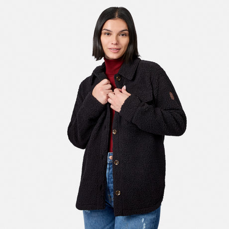 A woman with straight dark hair is standing against a plain white background, smiling softly as she holds the Free Country Women’s Sherpa Shacket—an oversized black jacket—over a red turtleneck and blue jeans.