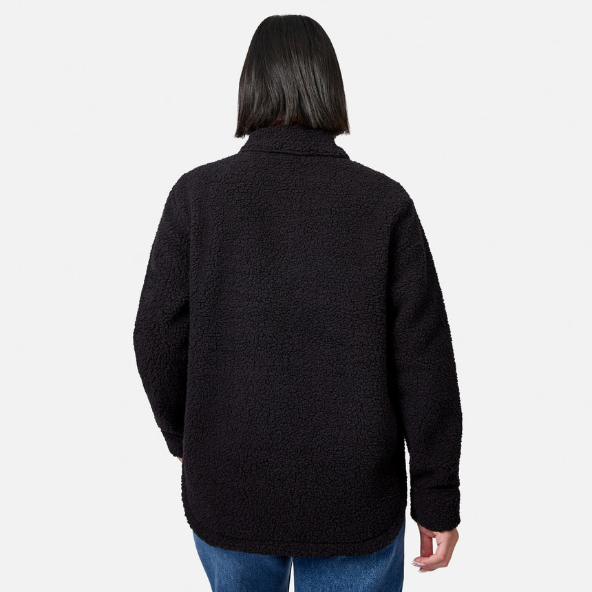 A woman with straight, shoulder-length dark hair is seen from behind, standing against a plain white background, wearing the Free Country Women’s Sherpa Shacket in black and blue jeans.
