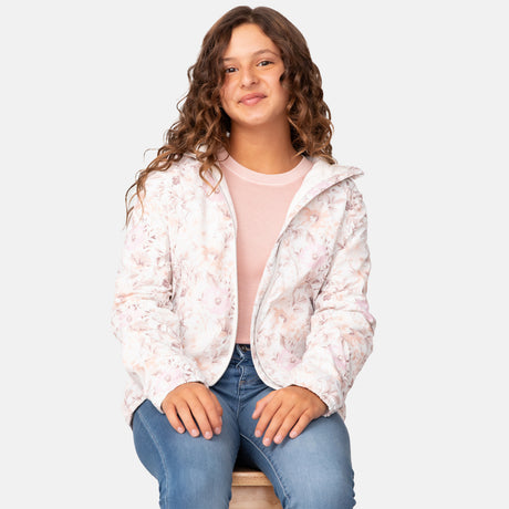 A young woman with long, curly brown hair sits on a stool, smiling. She is wearing the Clearance Girls' Windshear Jacket in light pink floral over a pink top and blue jeans, hands resting on her knees against a plain white background.