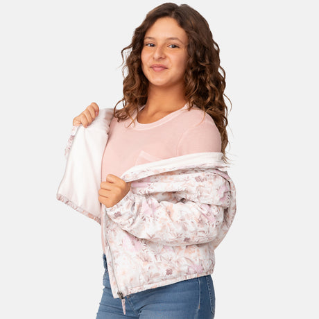 A smiling young woman with long curly hair wears a light pink shirt and blue jeans, putting on the Clearance Girls' Windshear Jacket—floral-patterned, wind and water resistant, and made from recycled polyester—against a white background.