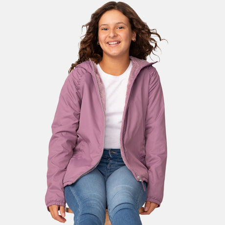 A smiling young person with wavy brown hair sits on a stool, wearing the Clearance Girls' Windshear Jacket—light purple, wind and water resistant—over a white shirt and blue jeans, against a plain light background.