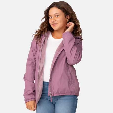 A young woman with long, wavy brown hair smiles and touches her hair against a plain background, wearing the Clearance Girls' Windshear Jacket in light purple over a white shirt and blue jeans.
