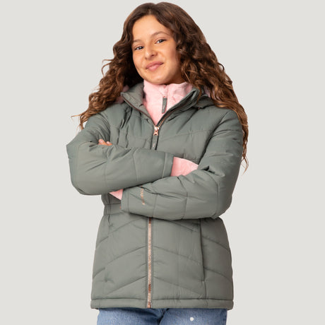 A young girl with long curly hair smiles with her arms crossed, wearing a Free Country Girls' Bib Puffer Jacket made from recycled polyester over a pink top and blue jeans, standing against a plain light gray background.