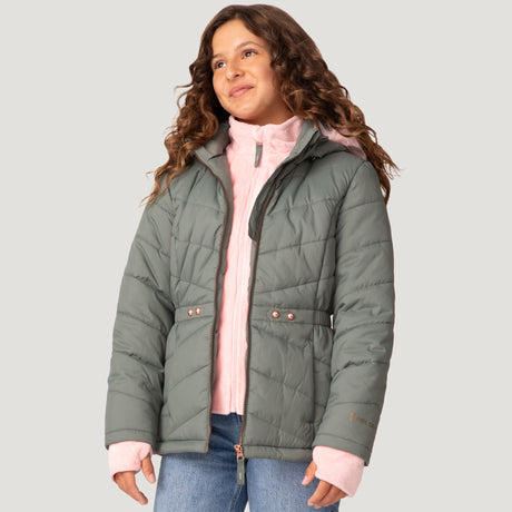 A girl with wavy brown hair wears the Free Country Girls' Bib Puffer Jacket over a light pink zip-up hoodie and blue jeans, smiling and looking slightly upward against a plain light background.