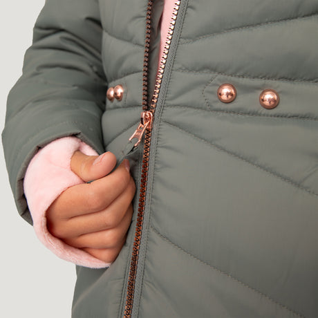 Close-up of a child’s hand zipping a water-resistant gray Girls' Bib Puffer Jacket by Free Country, featuring rose gold studs and zipper, with a light pink fuzzy sweater sleeve visible underneath. Made from recycled polyester.