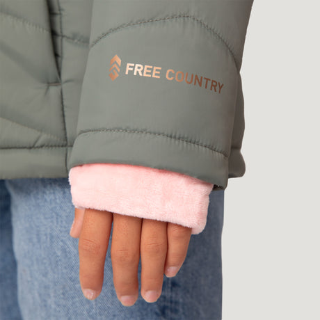 A close-up of a hand in a sage green Girls' Bib Puffer Jacket by Free Country, made with recycled polyester, water-resistant, "FREE COUNTRY" on the sleeve, pink fuzzy cuff underneath, and blue jeans visible.