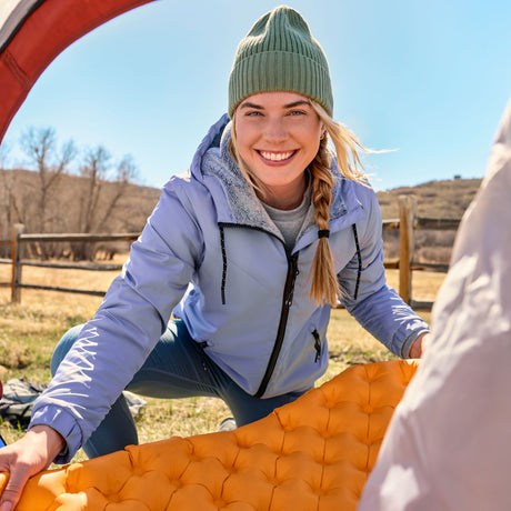Woman in blue Free Country windbreaker in outdoor setting with orange sleeping pad and blue sky