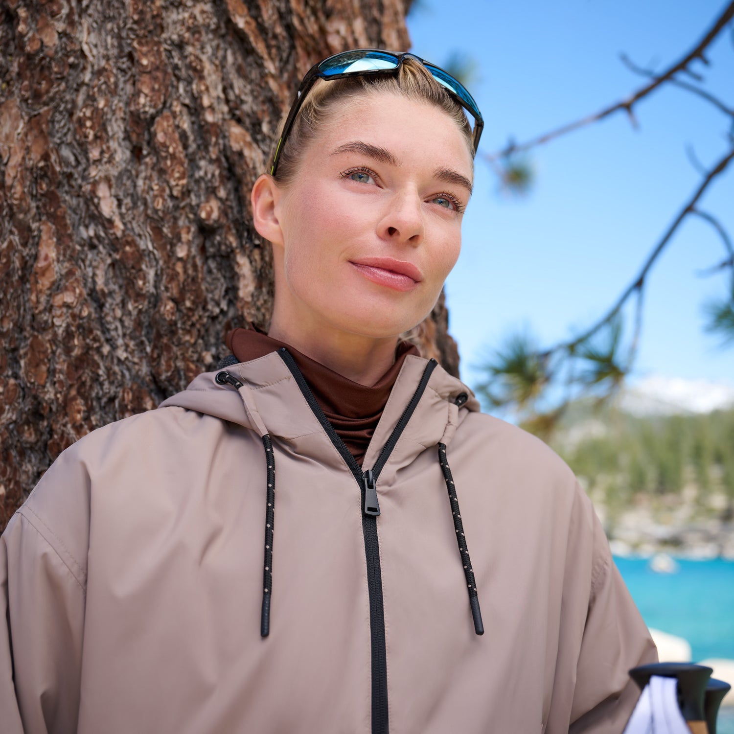 A woman in a tan Free Country Women’s Bomber Windshear Jacket and sunglasses leans against a tree outdoors, surrounded by pine branches and blue sky, enjoying a peaceful, sunny day near the water.