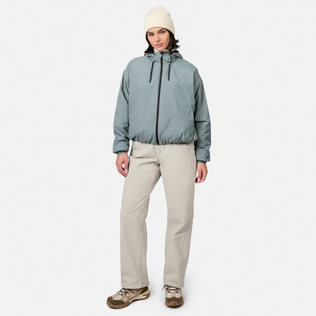 A person wears a light blue Free Country Women’s Bomber Windshear Jacket, beige pants, brown shoes, and a white beanie, standing against a plain white background facing the camera with one hand in their pocket.