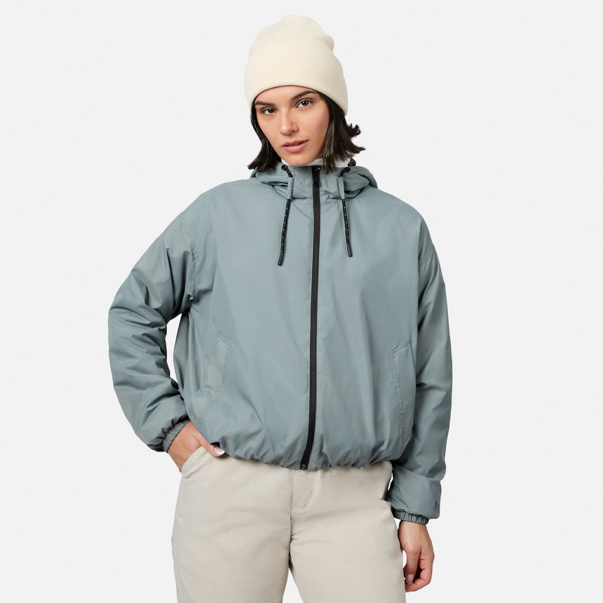 A person wears a Free Country Women’s Bomber Windshear Jacket in Cyprus, beige pants, and a cream knit beanie, standing with one hand in their pocket against a plain white background.