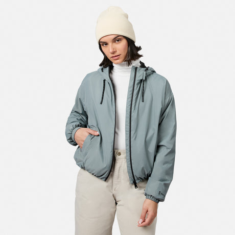 A woman models the Free Country Women’s Bomber Windshear Jacket in light gray, with a beige beanie, white turtleneck, and beige pants, standing against a plain white background with one hand in her pocket and a neutral expression.