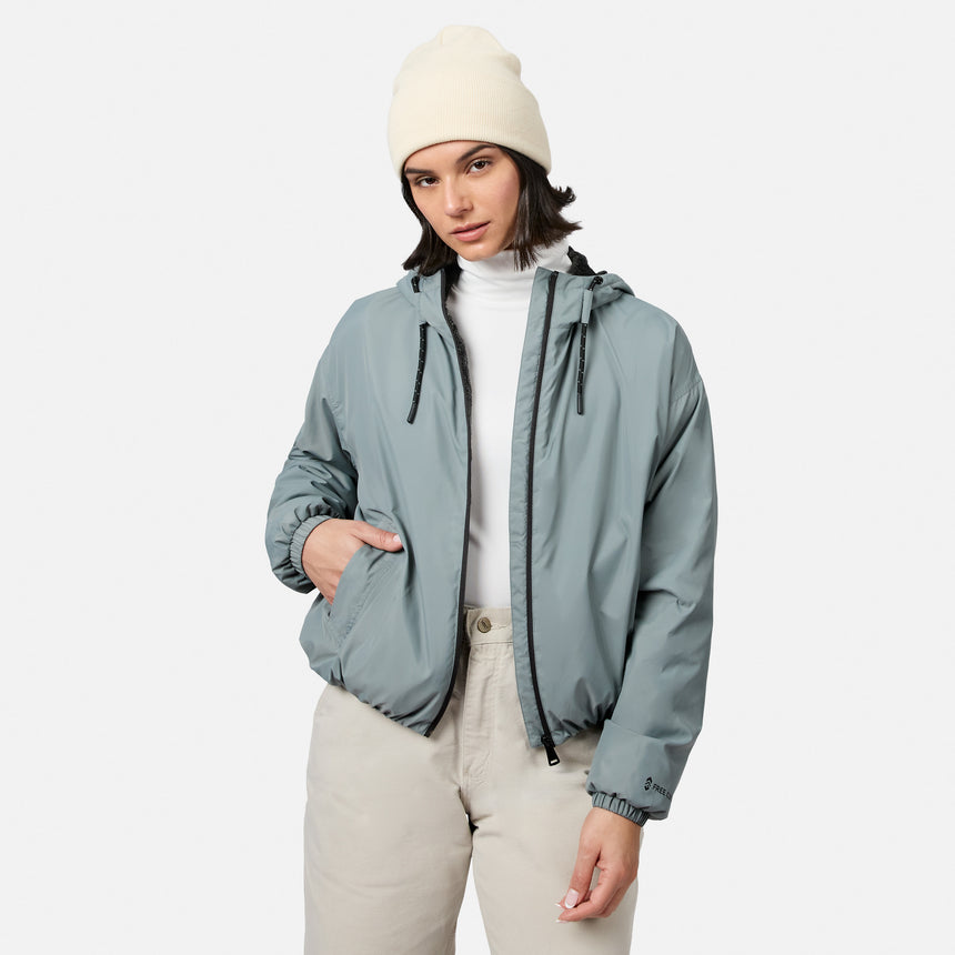 A woman models the Free Country Women’s Bomber Windshear Jacket in light gray, with a beige beanie, white turtleneck, and beige pants, standing against a plain white background with one hand in her pocket and a neutral expression.