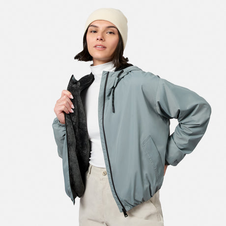 A woman wearing the Free Country Women’s Bomber Windshear Jacket and cream beanie stands against a plain background, opening her light gray jacket to reveal its black lining over a white turtleneck and beige pants.