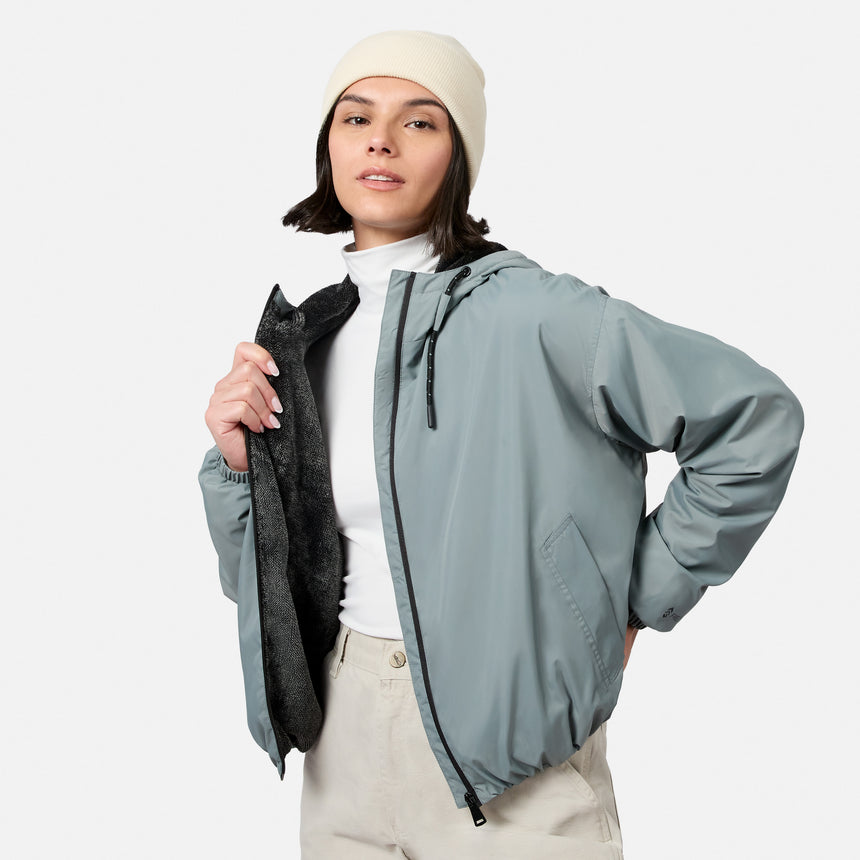 A woman wearing the Free Country Women’s Bomber Windshear Jacket and cream beanie stands against a plain background, opening her light gray jacket to reveal its black lining over a white turtleneck and beige pants.
