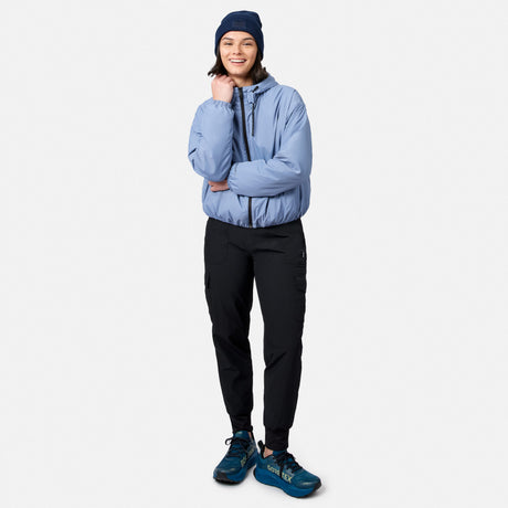 A smiling person wears the Free Country Women’s Bomber Windshear Jacket in light blue, with black pants, blue beanie, and blue sneakers. Against a white background, one arm is crossed while the other hand touches their chin.
