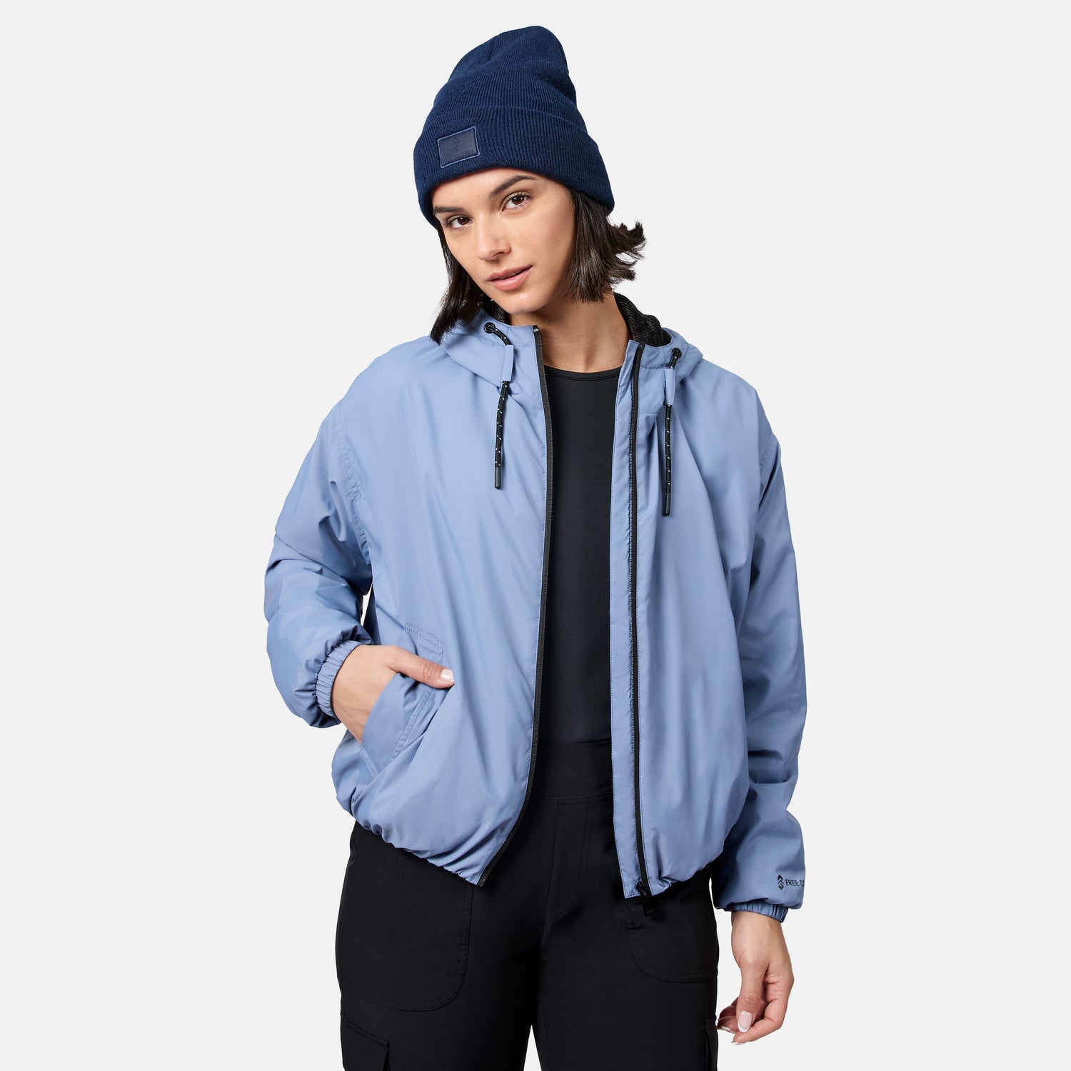 A woman models the Free Country Women’s Bomber Windshear Jacket in light blue, paired with a blue beanie and black pants, standing against a plain white backdrop with one hand in her pocket and a neutral look.