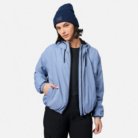 A woman models the Free Country Women’s Bomber Windshear Jacket in light blue, paired with a blue beanie and black pants, standing against a plain white backdrop with one hand in her pocket and a neutral look.