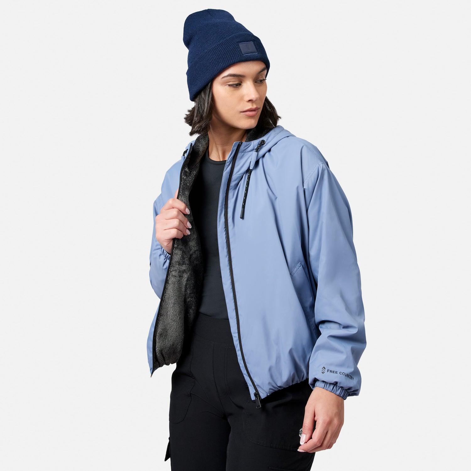 Model wears the Free Country Women’s Bomber Windshear Jacket in light blue with black lining, paired with a blue beanie and black pants. She looks left, holding the jacket open against a plain white background.