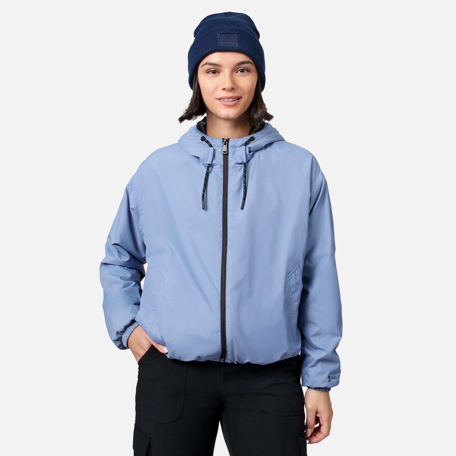A person wears the Free Country Women’s Bomber Windshear Jacket in Dusk, black pants, and a navy knit beanie, standing facing forward against a plain white background.