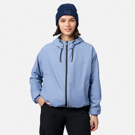 A person wears the Free Country Women’s Bomber Windshear Jacket in Dusk, black pants, and a navy knit beanie, standing facing forward against a plain white background.
