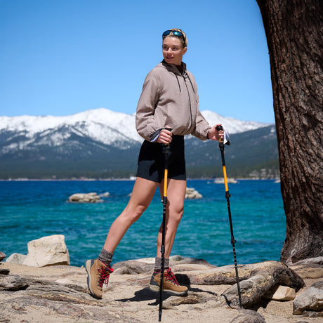 A hiker by a clear blue lake with snowy peaks, holding trekking poles and wearing boots, shorts, and a Women’s Bomber Windshear Jacket by Free Country on a sunny day.