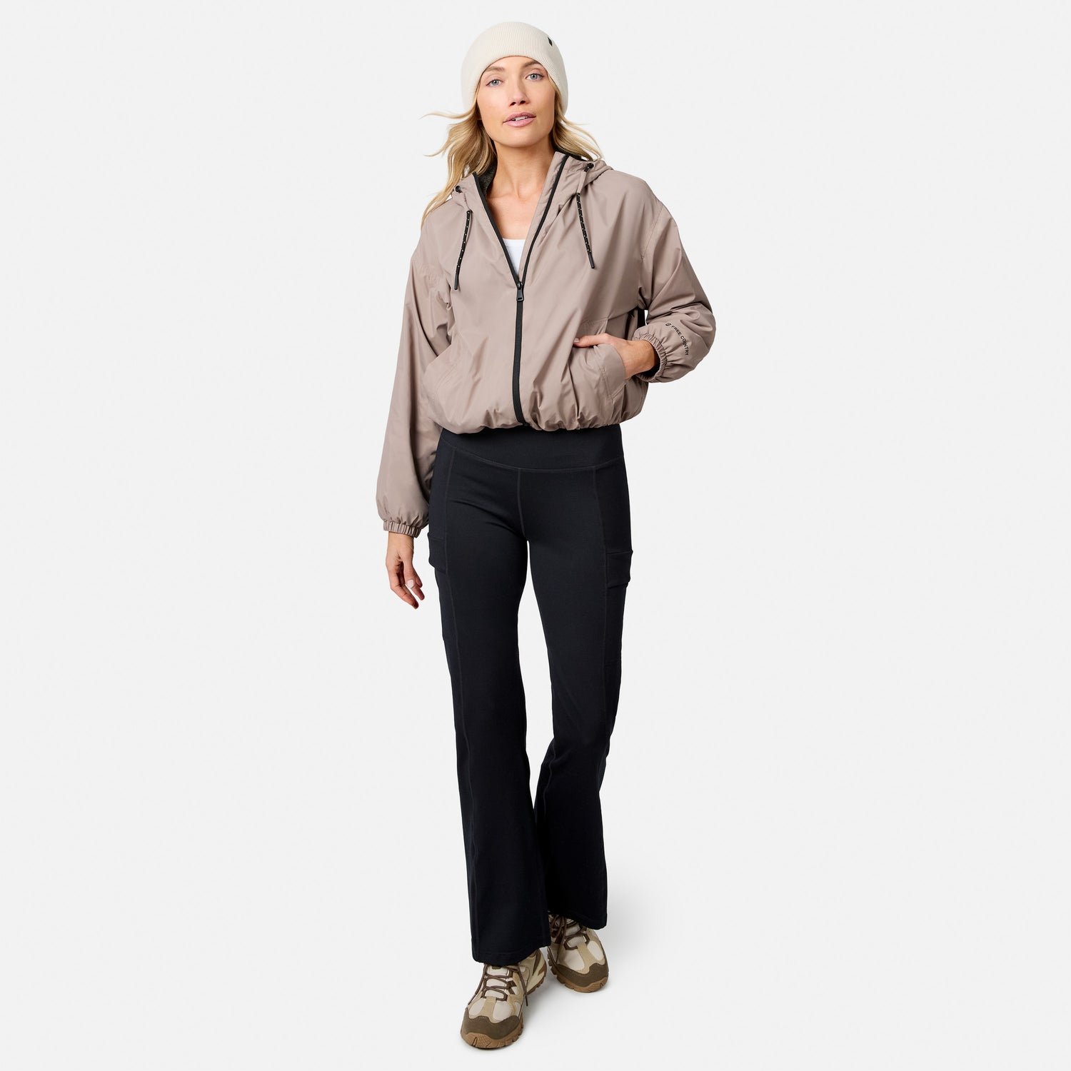 A woman wears a Free Country Women’s Bomber Windshear Jacket, beige beanie, white top, black flare pants, and tan sneakers while standing against a plain white background.