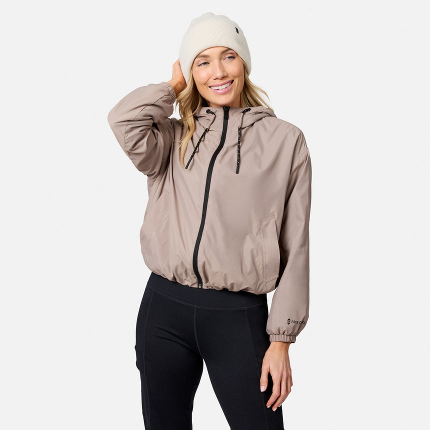 A woman in the Free Country Women’s Bomber Windshear Jacket, black leggings, and a white beanie smiles with one hand on her head, standing against a plain light background.