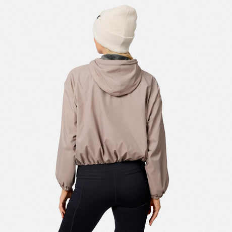 A person wearing a light beige beanie, black leggings, and the Free Country Women’s Bomber Windshear Jacket in taupe stands with their back to the camera against a plain white background.