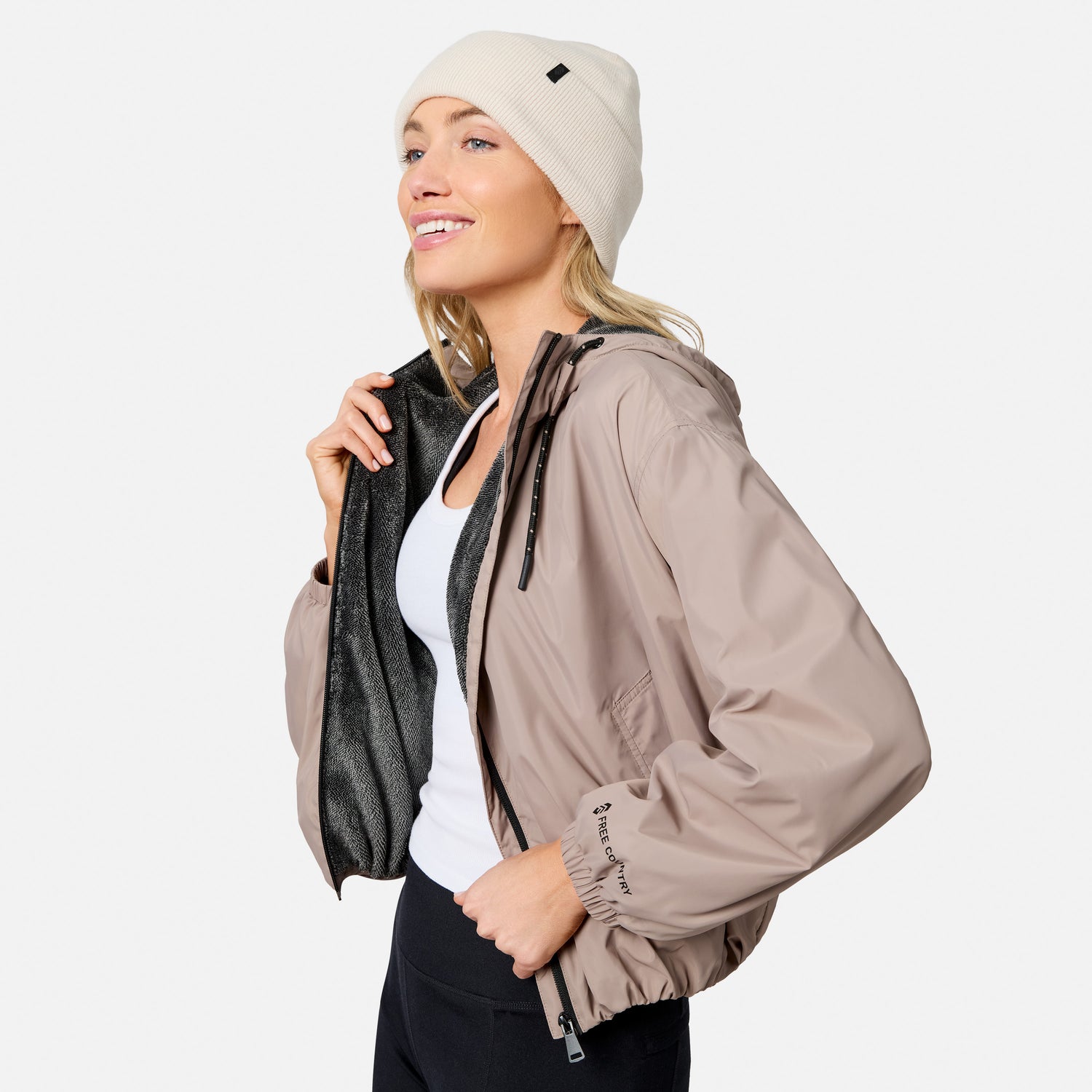 Smiling woman in a beige beanie, white top, black pants, and the Free Country Women’s Bomber Windshear Jacket—light brown with black lining—standing against a plain light background.