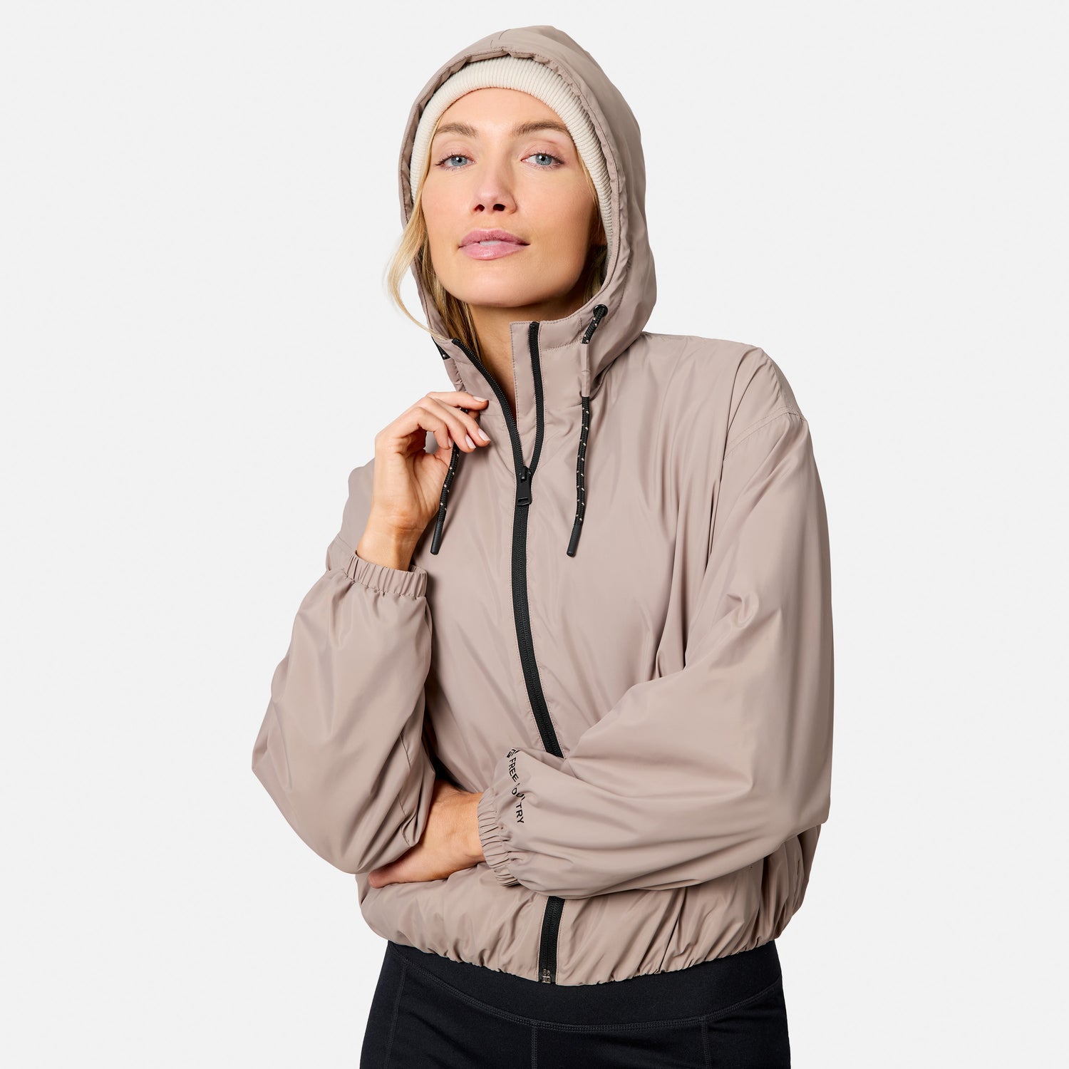 A woman models the Free Country Women’s Bomber Windshear Jacket in tan, made from recycled polyester, over a light beanie. She holds the collar and looks confidently at the camera against a plain background.