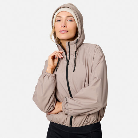 A woman models the Free Country Women’s Bomber Windshear Jacket in tan, made from recycled polyester, over a light beanie. She holds the collar and looks confidently at the camera against a plain background.