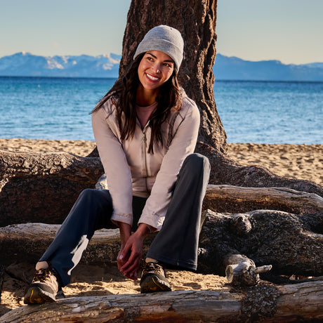 A woman in a gray beanie and Free Country Women’s Anorak Windshear Jacket smiles while sitting on driftwood at a sandy beach, with mountains and blue water in the background.
