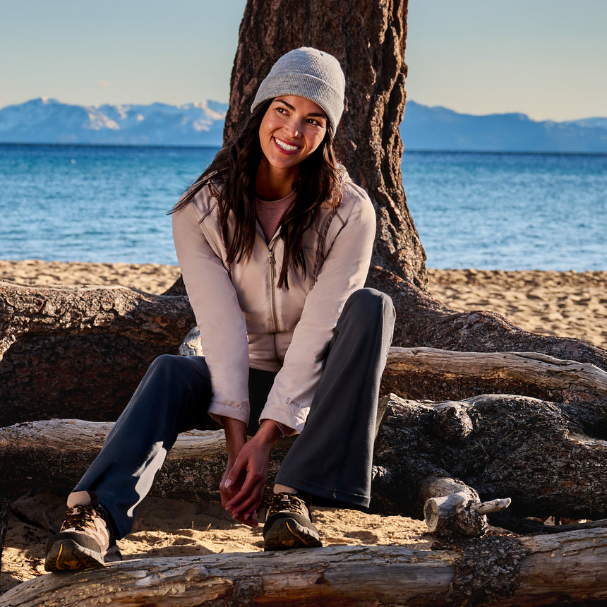 A woman in a gray beanie and Free Country Women’s Anorak Windshear Jacket smiles while sitting on driftwood at a sandy beach, with mountains and blue water in the background.