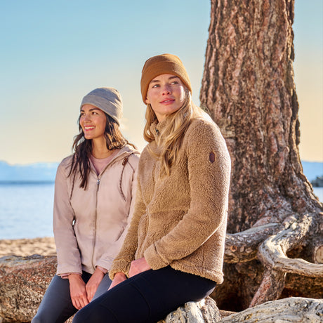 Two women wearing Free Country Women’s Sierra Butter Pile® Zip Up Jackets and knit hats sit on tree roots by a lake. Sunlight filters through the clear sky, making the outdoor scene bright and inviting.
