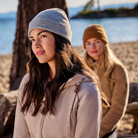 Two women wear Free Country Women’s Anorak Windshear Jackets and knit hats on a wintry beach. One stands gazing ahead, the other sits on a log. Trees and water frame the outdoor scene.