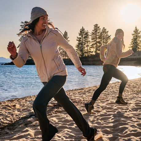 Two women wearing Free Country Women’s Anorak Windshear Jackets run and smile on a sandy beach at sunset near water, with pine trees silhouetted against the golden sky.