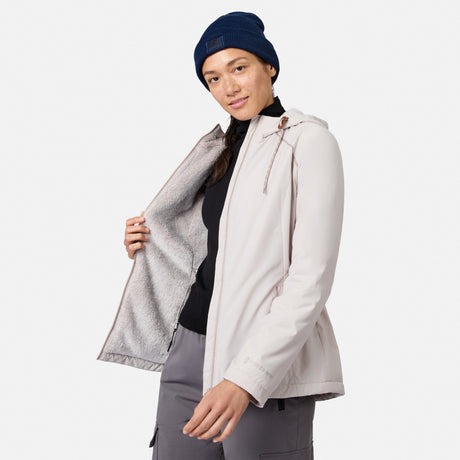 A woman in a light Women’s Anorak Windshear Jacket by Free Country, over a dark shirt, gray pants, and a blue beanie, holds open the jacket to reveal its soft lining. The background is plain white.