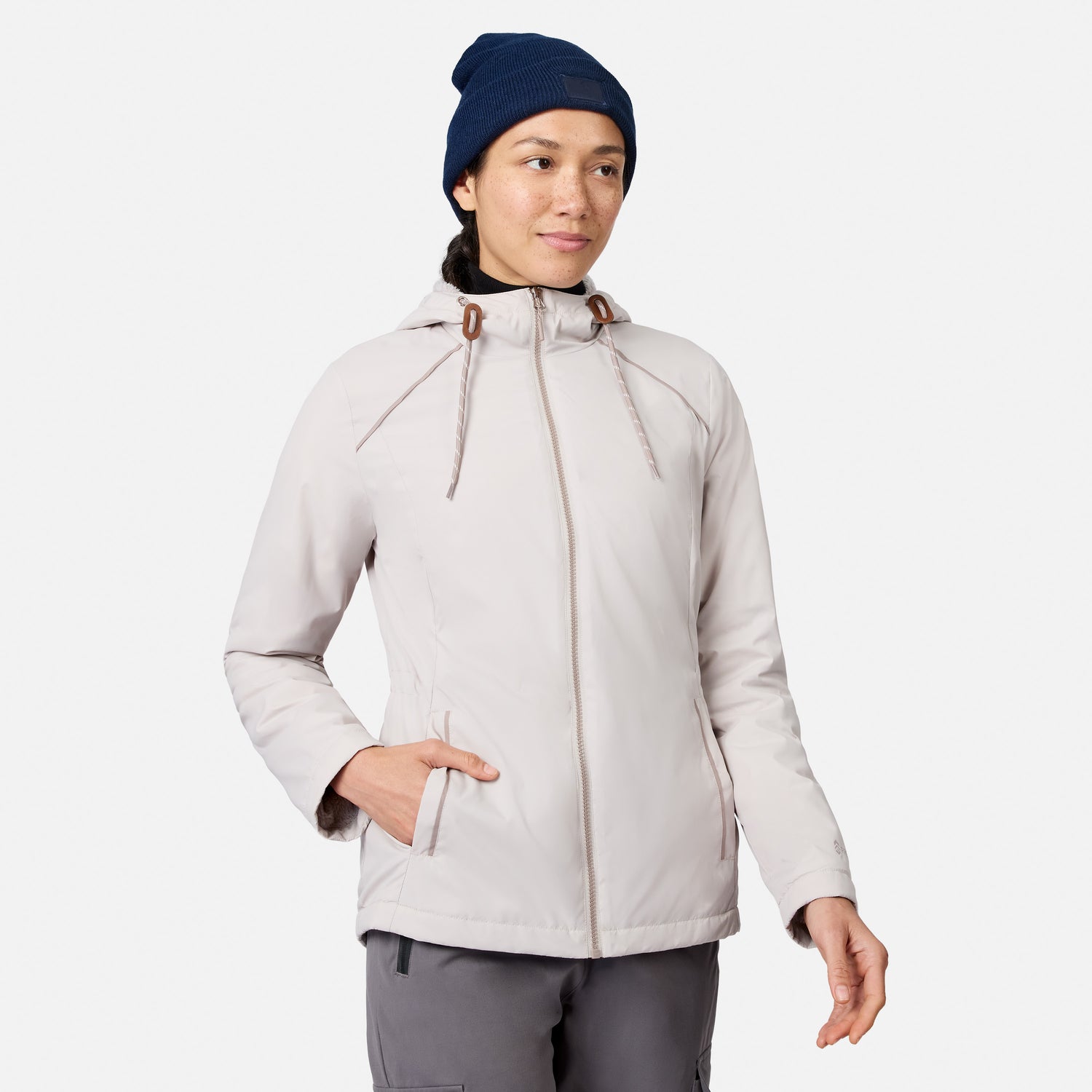 A person wearing a Women’s Anorak Windshear Jacket by Free Country in bisque stands against a white background with one hand in their pocket, looking slightly to the side and smiling softly.