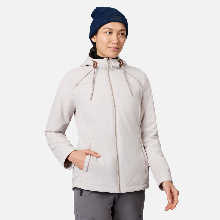 A person wearing a Women’s Anorak Windshear Jacket by Free Country in bisque stands against a white background with one hand in their pocket, looking slightly to the side and smiling softly.