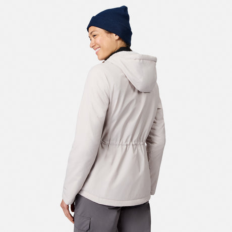 A woman in a Free Country Women’s Anorak Windshear Jacket with hood, navy beanie, and gray pants stands facing away, smiling over her shoulder against a plain white background.