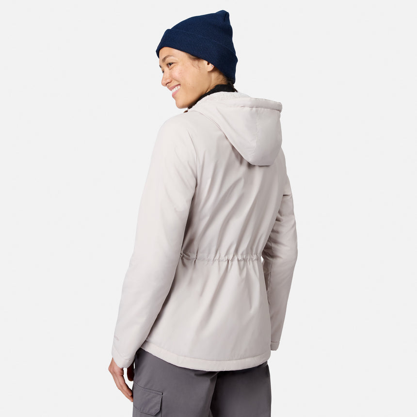 A woman in a Free Country Women’s Anorak Windshear Jacket with hood, navy beanie, and gray pants stands facing away, smiling over her shoulder against a plain white background.