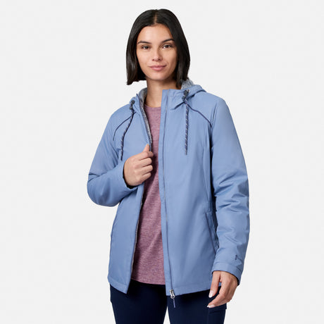 A woman with shoulder-length dark hair models the Free Country Women’s Anorak Windshear Jacket in light blue over a mauve shirt and dark pants, standing against a plain white background, facing forward and holding the jacket partially open.