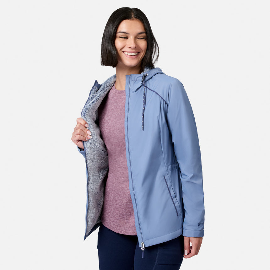 Smiling woman with a bob haircut models the Free Country Women’s Anorak Windshear Jacket in light blue over a pink shirt, showing off its soft, textured lining against a plain white background.