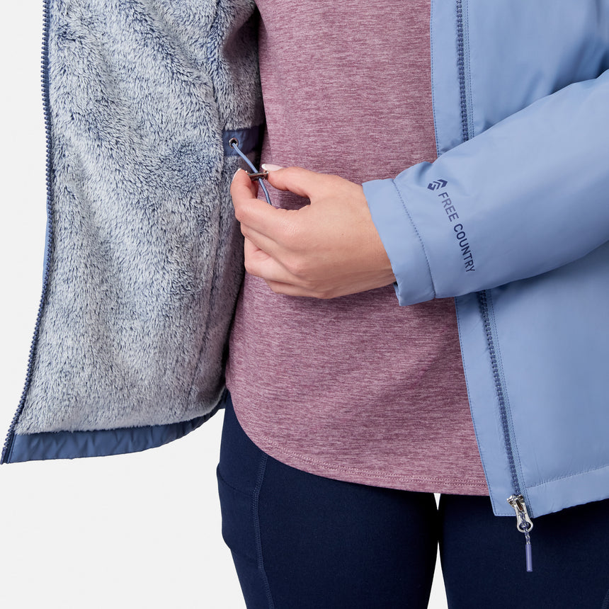 A person in a Free Country Women’s Anorak Windshear Jacket—light blue, water-resistant, with fuzzy grey lining—zips an inner pocket; mauve shirt and dark blue pants visible. Only torso and hand shown against a white background.